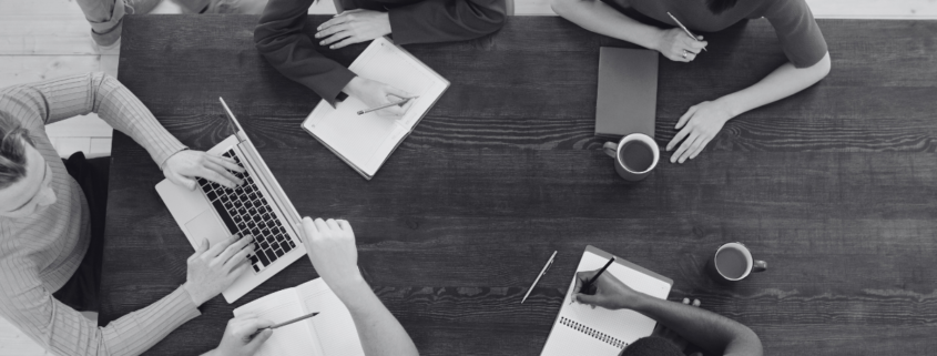 colleagues meeting in an office around a dark wood table with notepads and a laptop